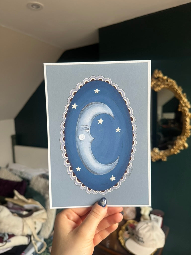 Hand holding a card with a blue crescent moon and stars design against a light blue background.